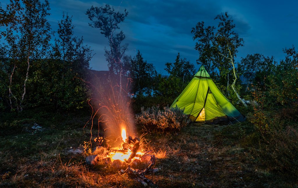 von innen beleutetes Zelt mit Lagerfeuer im Vordergrund vor Abendhimmel