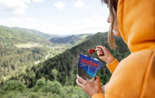 Junge Frau isst gefriergetrocknete Lebensmittel zum Wandern aus Spezialverpackungen und genießt die großartige Landschaft, während sie hoch in den Bergen unterwegs ist